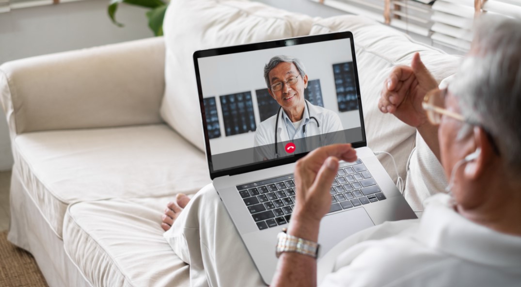 Senior sitting on couch with laptop open with a male doctor appearing on screen during telehealth call